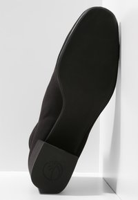 Suela de zapato negro de punta cerrada con tacón bajo,底 plana y logo circular grabado en el tacón, inclinada contra una pared y suelo blancos.