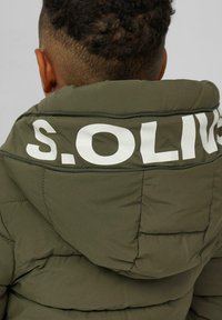 Kind mit kurzen lockigen Haaren, das eine olivgrüne Steppjacke mit dem Aufdruck "S.OLIVER" auf der Kapuze trägt, von hinten gesehen.