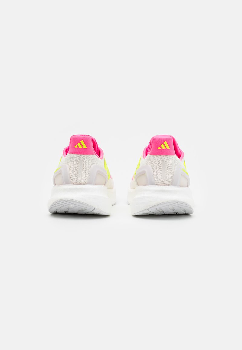 Par de zapatillas deportivas blancas con tacones rosas, rayas amarillas neón y suelas texturizadas blancas, vistas desde atrás sobre un fondo blanco.