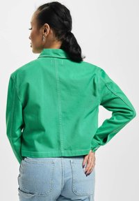 Mujer con coleta negra que lleva una chaqueta verde corta y unos vaqueros azules de talle alto, con la mano apoyada en el bolsillo trasero.