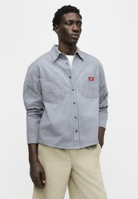 Άνδρας που φοράει μπλε και λευκό πουκάμισο με κατακόρυφες ρίγες και τσέπες στο στήθος, καθώς και μπεζ παντελόνι, στέκεται με το ένα χέρι στην τσέπη.