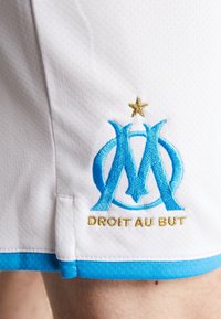 Shorts de sport blancs avec une bordure bleue. Présente un logo bleu brodé et le texte doré "DROIT AU BUT" accompagné d'une étoile dorée au-dessus du logo.