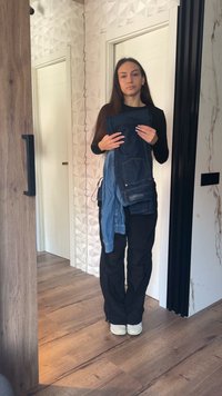 Νεαρή γυναίκα με μαύρο ντύσιμο κρατά διπλωμένα τζιν ρούχα, στέκεται σε έναν σύγχρονο χώρο με ξύλινο δάπεδο και λευκό τοίχο με υφή.