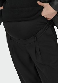 Mustat äitiysleggingsit, joissa on sileä pinta, korkeavyötäröinen ja edessä laskosyksityiskohta. Suunnittelussa on säädettävät sivunauhat.