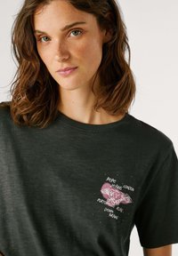 T-shirt in cotone grigio scuro con maniche corte, caratterizzato da una grafica rosa di un cuore con ali e testo. Tessuto morbido e vestibilità rilassata.