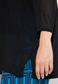 Schwarzes Oberteil mit einem strukturierten Muster und seitlichem Schlitz, kombiniert mit blauen gemusterten Hosen, die das komplizierte Design und den Farbkontrast betonen.