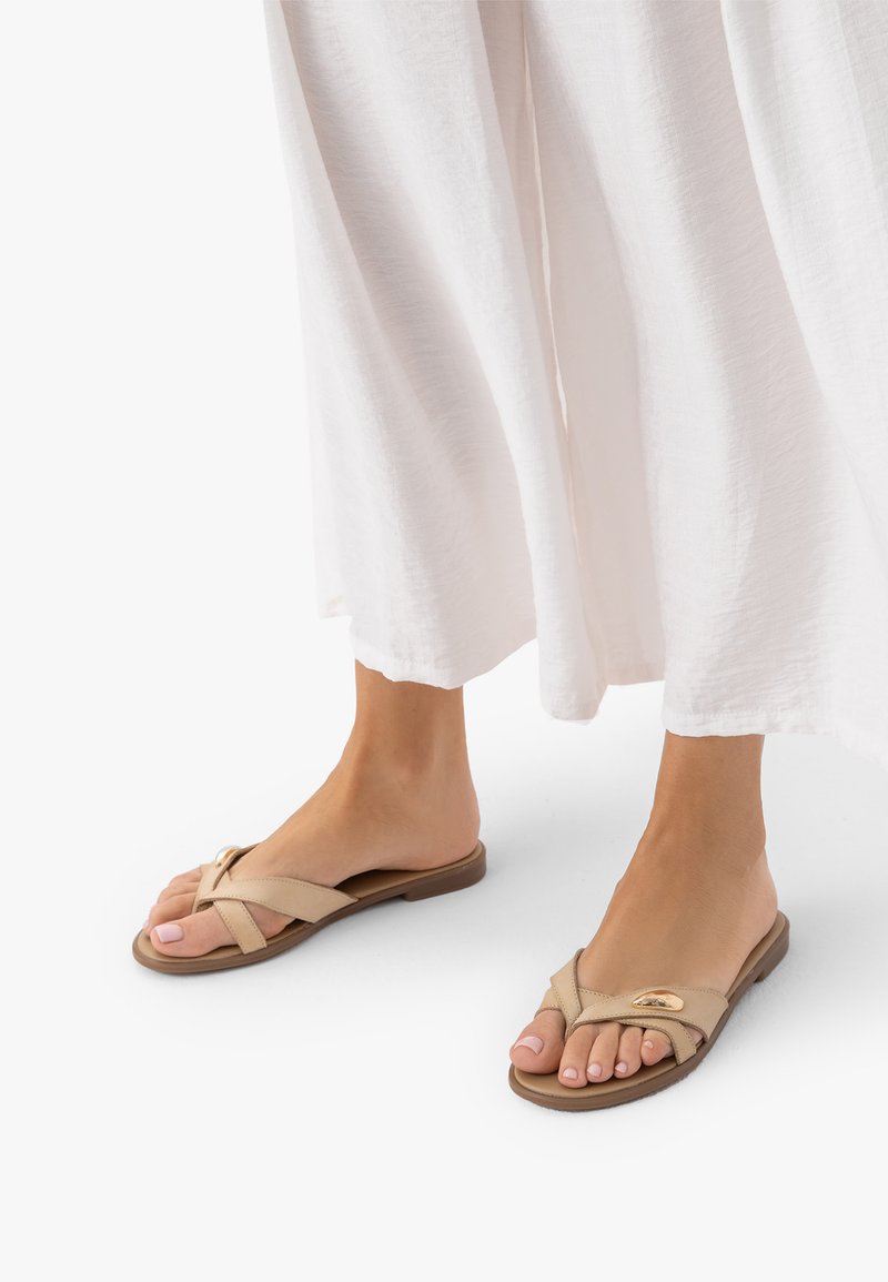 Pieds portant des sandales plates beiges à lanières croisées avec un détail doré, associées à une jupe blanche fluide longueur cheville.