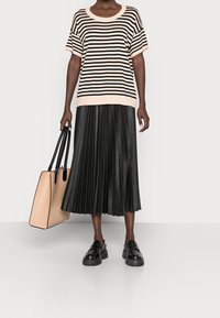 Vrouw in een beige en zwarte gestreepte top, zwarte geplooide midi-rook, zwarte chunky loafers en met een beige tote bag met zwarte strap.