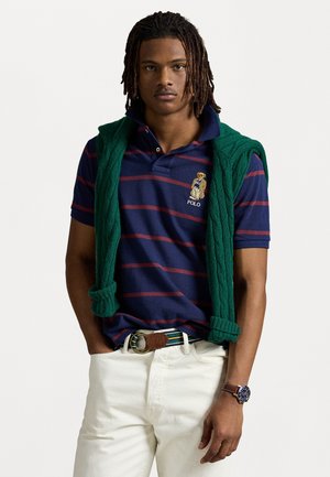 Marine gestreepte polo met een groene kabelgebreide trui over de schouders. Crème kleurige broek en een multikleurige riem met een leren gesp. Zilveren horloge.