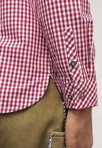 Chemise à carreaux rouges et blancs en coton, avec poignets boutonnés et ourlet arrondi. Le tissu a une texture lisse avec des coutures visibles.