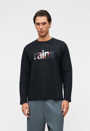 Young man wearing a black long-sleeve shirt with "rains" printed in gradient letters, paired with loose gray pants against a plain background.