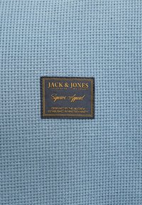 Μπλε ύφασμα πλεκτού βαφής με μια μαύρη και χρυσή ετικέτα ρούχων Jack & Jones ραμμένη στο κέντρο.