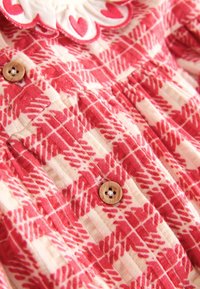 Rdeč in bel vzorčast material z detajli gumbov in obrobo z valovitimi robovi, ki prikazuje naborke na oblačilu.