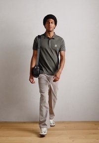 Polo verde măsline cu un logo aplicat, pantaloni bej deschis și adidași albi. Geantă de umăr neagră cu un design texturat. Fundal din podea din lemn.