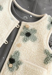 Chaleco de forro polar en crema con patrones florales verdes, con un ribete beige, dos botones y un bolsillo. Debajo, se ve una camiseta gris de canalé.
