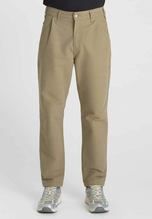 Man die khaki rechte broek en zilvergrijze hardloopschoenen draagt, staat tegen een effen lichte achtergrond.