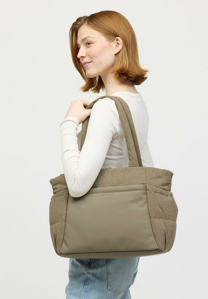 Junge Frau mit rotem Haar, die ein weißes Oberteil und Jeans trägt, eine große beige Tragetasche über der Schulter trägt und seitlich schaut.