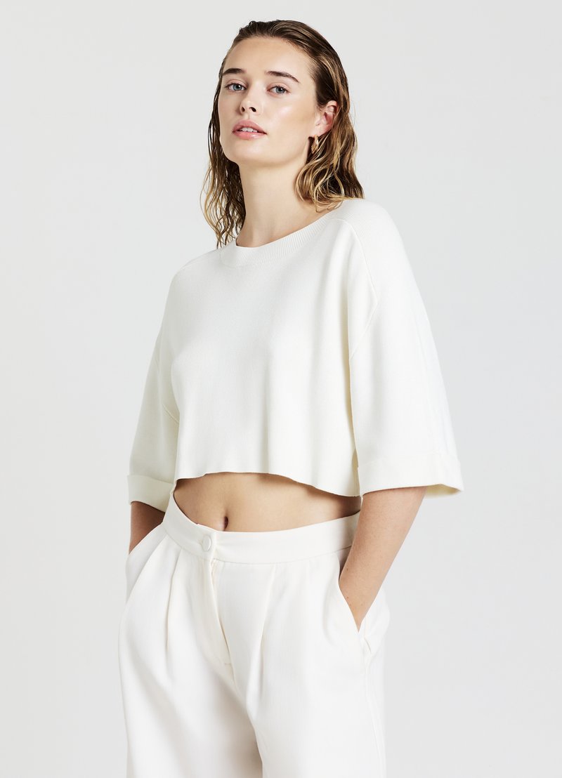 Maglione bianco corto con una texture a coste, maniche ampie e scollatura rotonda, abbinato a pantaloni bianchi a vita alta con pieghe.