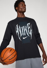 Schwarzes Langarm-Nike-Shirt mit einem großen Flammenlogo in Silber und Blau auf einem Gitterhintergrund. Graue Sportshorts und ein oranger Basketball.