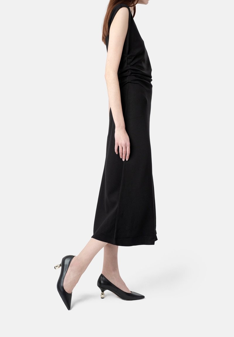 Femme portant une robe midi noire sans manches et des talons noirs pointus avec des talons métalliques ronds dorés uniques, debout sur une jambe.