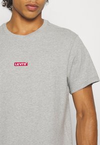 Tricou din bumbac gri, cu un logo roșu rectangular Levi's. Are mâneci scurte și un gât rotund, având o textură netedă.