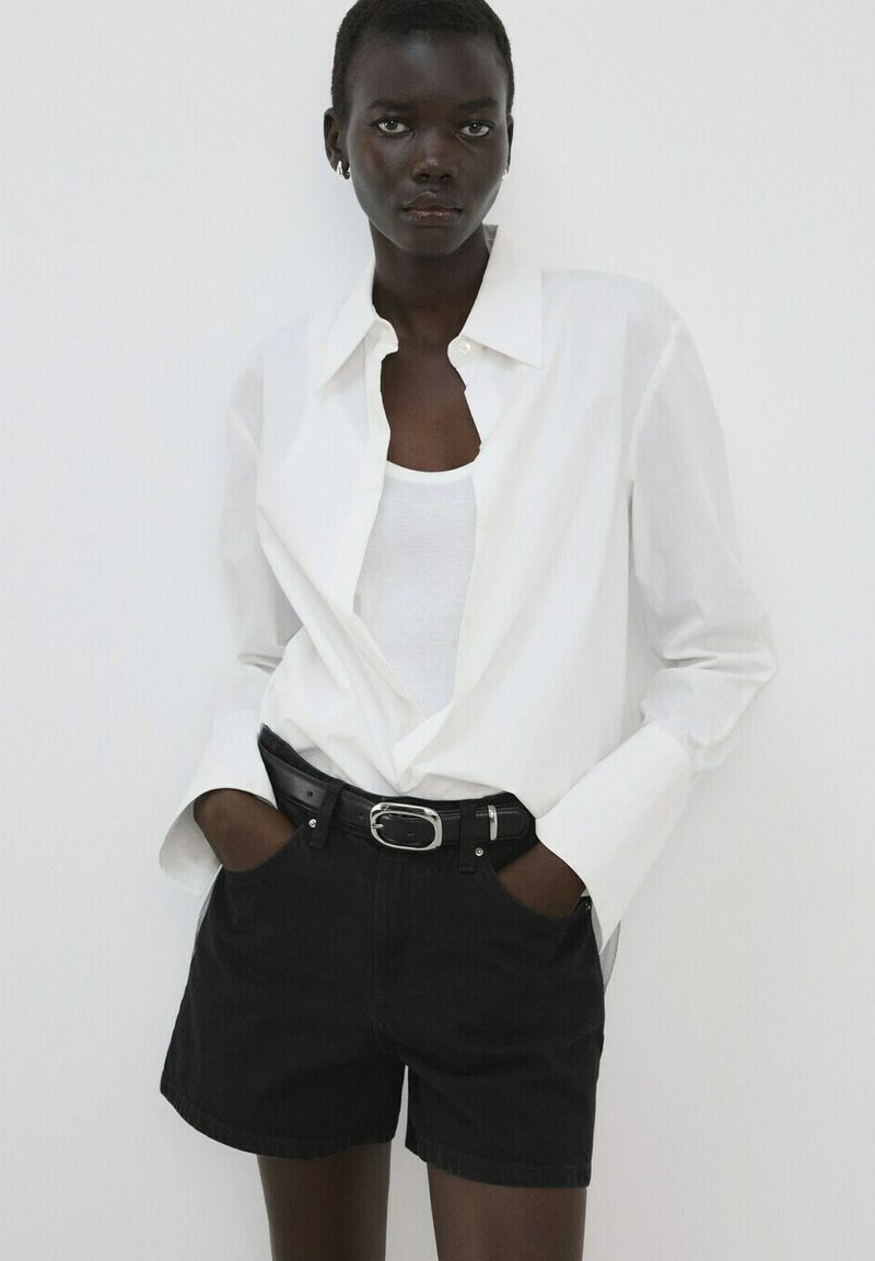 Femme portant une chemise blanche boutonnée par-dessus un débardeur blanc, une ceinture noire et un short noir, debout les mains dans les poches devant un fond blanc.