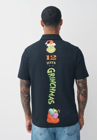 Musta lyhythihainen paita, jossa värikkäitä Grinch-kuvioita, teksti "12 Days of Grinchmas" sekä lahjapaketti, jossa on käärittyjä lahjoja ja karamellitankoja.