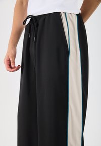 Melnas sporta bikses ar aukliņu jostasvietā, ar sānu paneļiem beidža un zilo akcentu krāsā. Gluda tekstūra un brīva piegriezuma stils.