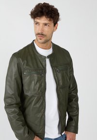 Olivenfarbene Lederjacke mit rundem Kragen, zwei Reißverschlusstaschen auf der Brust, glatter Textur und durchgehenden Reißverschluss, getragen über einem weißen Hemd.