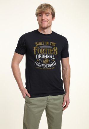 Homme aux cheveux châtain clair portant un t-shirt noir avec l'inscription "Built in the Forties Original and Unrestored" et un pantalon vert, debout les mains dans les poches.