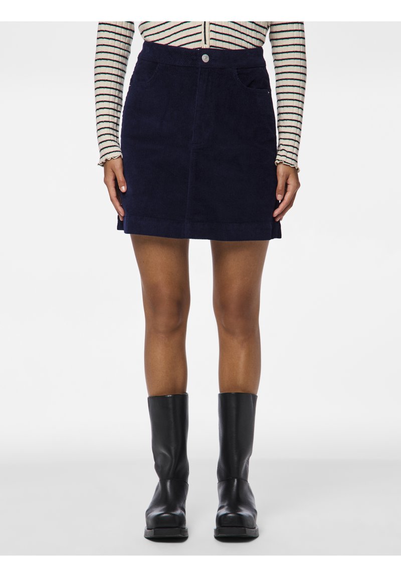 Femme portant une mini-jupe en velours côtelé bleu marine, des bottes mi-mollet noires et une chemise rayée à manches longues, debout devant un fond uni.