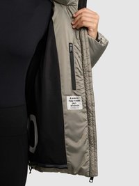 Polsterēta jaka gaiši pelēkā krāsā ar melnu oderi, kurai ir rāvējslēdzēja kabata un etiķete ar uzrakstu "Silts apskāviens no auduma."