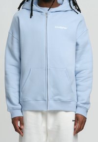 Sweat à capuche zippé bleu clair, fabriqué en tissu doux. Comprend une capuche, des poches avant, des poignets côtelés et un petit logo sur la poitrine.