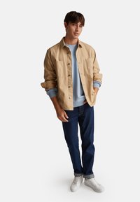 Chaqueta de botón en tonos beige sobre sudadera azul claro, jeans oscuros y zapatillas blancas. Corte ajustado con dos bolsillos en el pecho y mangas remangadas.
