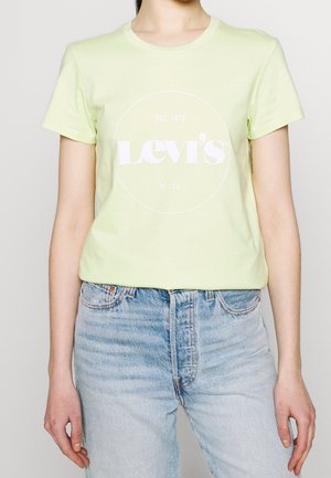Person iført en bleg gul Levi’s T-shirt med hvidt logo og lyseblå high-waisted jeans, stående mod en ensfarvet baggrund.