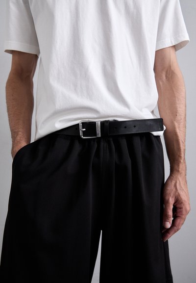 Black leather belt with a silver rectangular buckle, worn over black pleated trousers, paired with a plain white t-shirt.