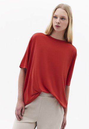 Ung kvinde med langt blondt hår iført løstsiddende rød top med korte ærmer og beige bukser, stående foran en ensfarvet hvid baggrund.