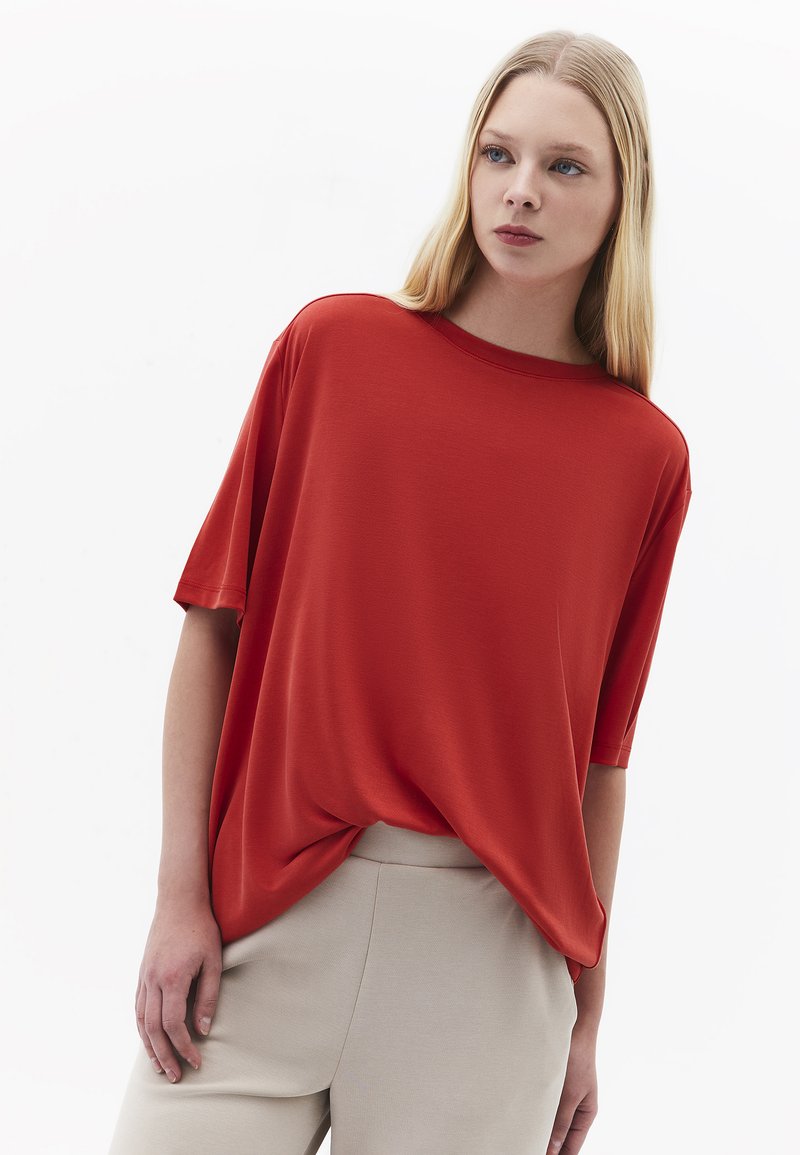 Jonge vrouw met lang blond haar draagt een losse rode top met korte mouwen en beige broek, staand tegen een effen witte achtergrond.