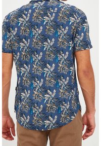 Koszula z krótkim rękawem w niebieskim materiale z tropikalnym wzorem liści. Posiada kontrastujące białe i zielone elementy oraz zaokrąglony dół.