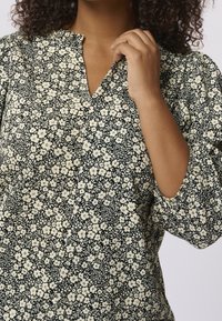 Czarna i kremowa bluzka w kwiaty z dekoltem w serek, bufiastymi rękawami i teksturowanym materiałem. Posiada mały wzór kwiatowy na ciemnym tle.