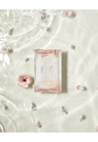 L'emballage du masque en gel de pivoine présente un doux dégradé de rose. Il est placé sur de l'eau parsemée de pétales de fleurs roses pâles, créant une composition épurée et sereine.