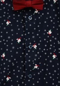 Mornarica majica s uzorkom pahuljica i polarnog medvjeda, crvena leptir-mašna, plavi gumbi. Mekana tkanina, detaljan print i simetrično postavljanje gumba.