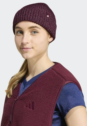 Jonge vrouw met blond haar draagt een bordeauxkleurige gebreide muts en bijpassend bordeauxkleurig fleecevest over een blauw shirt, met een lichte glimlach naar voren kijkend.
