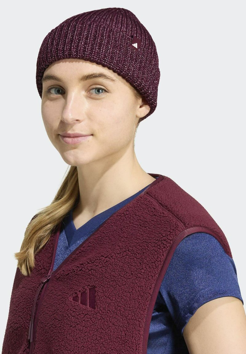 Jeune femme aux cheveux blonds porte un bonnet en tricot bordeaux et une veste polaire bordeaux assortie par-dessus une chemise bleue, faisant face avec un léger sourire.