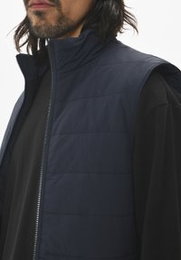 Navy puffer vest with horizontal quilting, high collar, and front zipper, layered over a black long-sleeve shirt, showcasing a smooth texture.