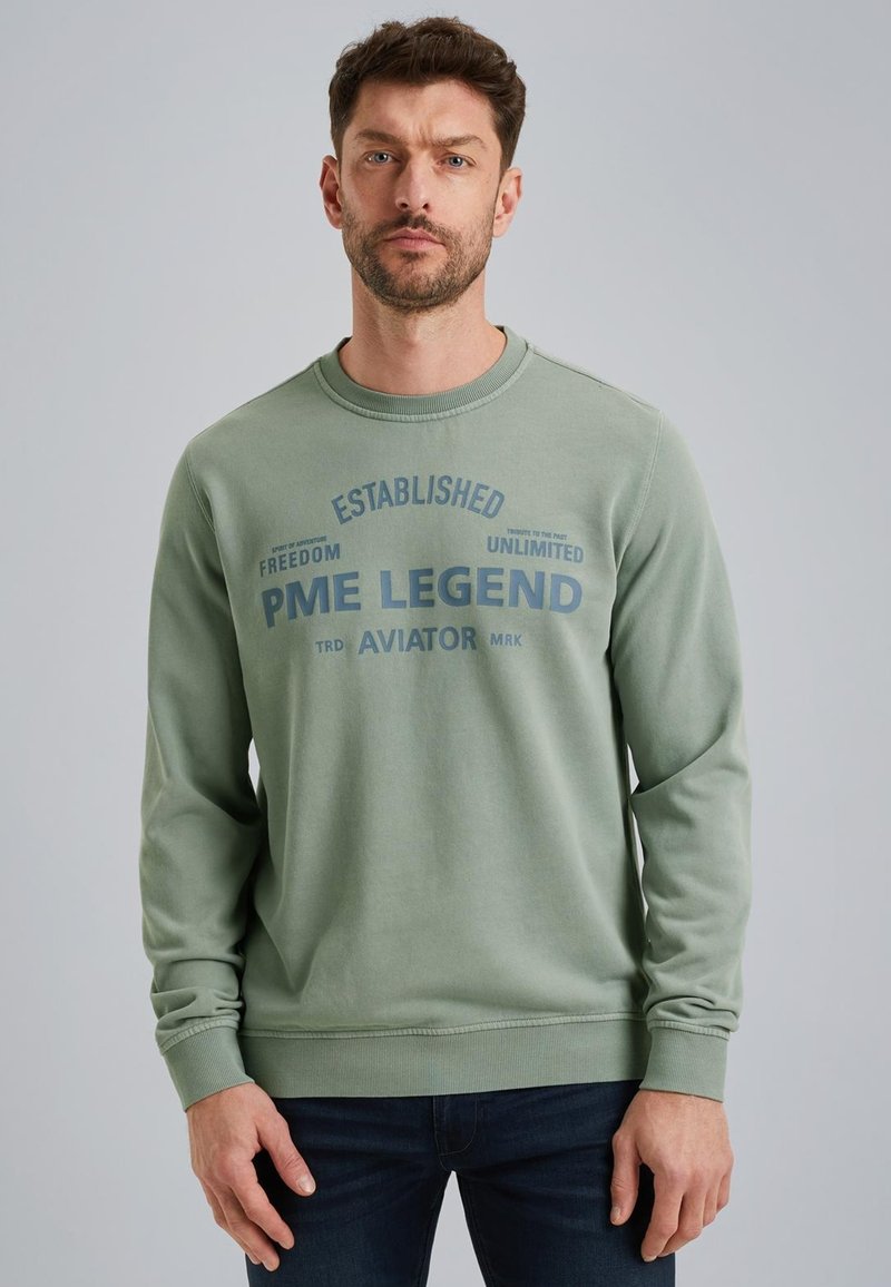 Sudadera verde de una mezcla suave de algodón con cuello redondo, mangas largas y un estampado gráfico central con texto en azul más oscuro.