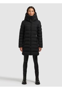 Kvinde iført en lang sort foret vinterjakke med høj krave, sorte leggings og sorte støvler, stående foran en ensartet lys baggrund.