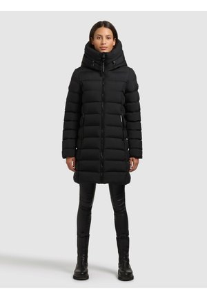 Frau trägt einen langen schwarzen gesteppte Wintermantel mit hohem Kragen, schwarze Leggings und schwarze Stiefel, steht vor einem einfarbigen hellen Hintergrund.