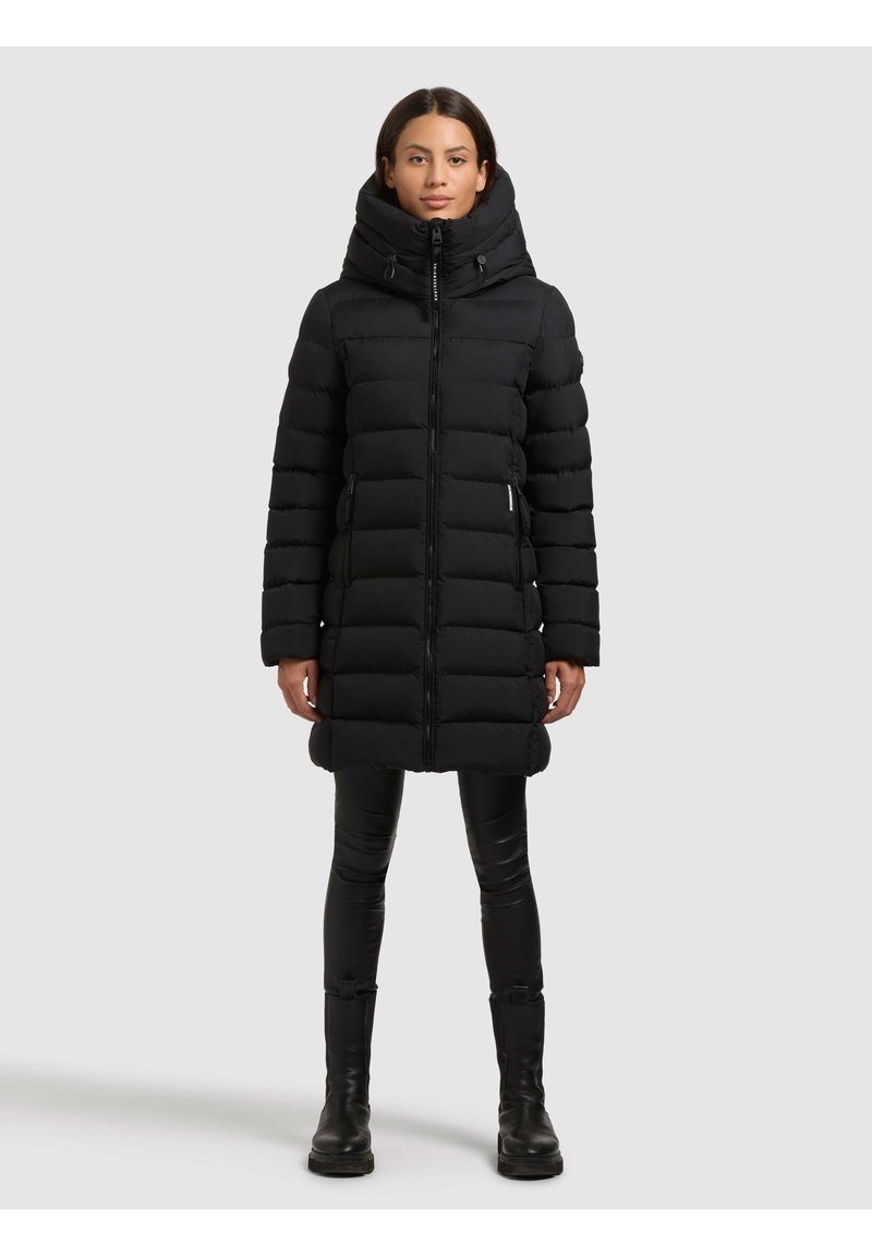 Kvinde iført en lang sort foret vinterjakke med høj krave, sorte leggings og sorte støvler, stående foran en ensartet lys baggrund.