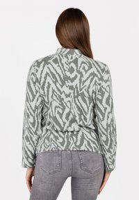 Polarowy sweter w subtelnym zielono-białym wzorze zebry, z długimi rękawami i kołnierzem typu szal, w połączeniu z szarymi dżinsami.